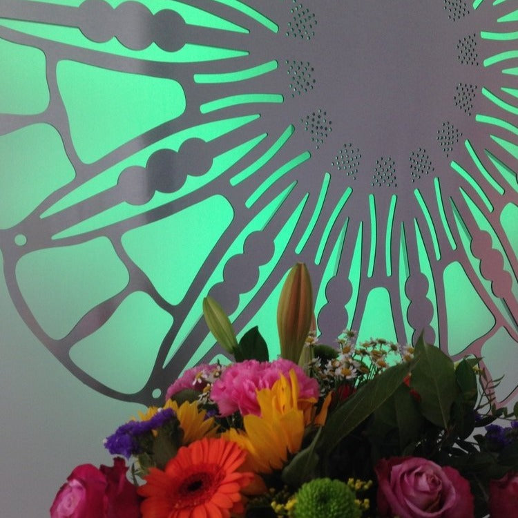 Close-up of KuvaLight Anemone wall lamp (115 cm, brushed aluminum) with green light, complementing a bouquet of flowers.