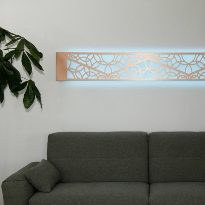 KuvaLight Minho 1 in brushed copper hanging horizontally above a couch with a light blue color light. Photo taken in a home setting.